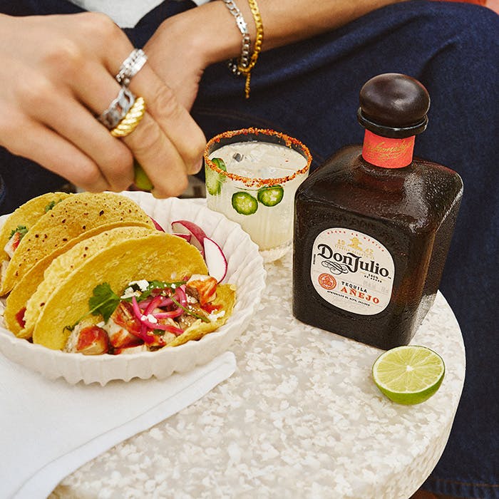 A bottle of Don Julio Anjeo tequila on a table next to a spicy margarita cocktail and tacos