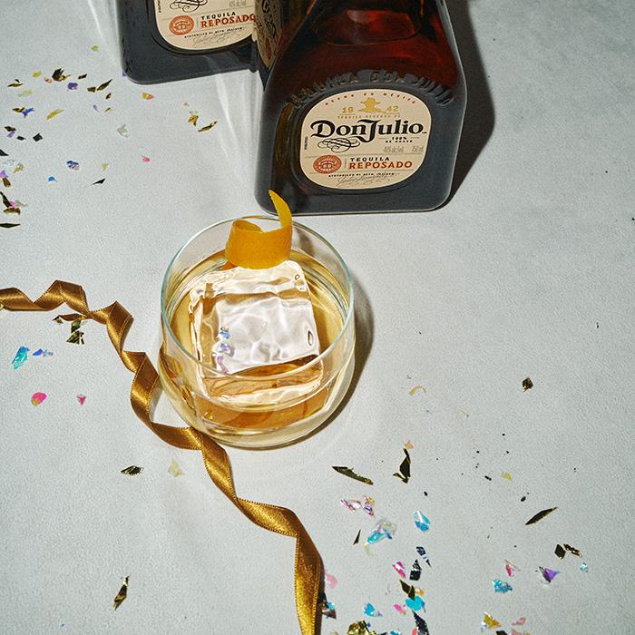 A bottle of Don Julio Reposado tequila next to an Old Fashioned cocktail and party ribbons