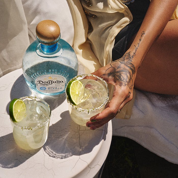 A bottle of Don Julio Blanco tequila on a table next to two margarita cocktails. A person's hand is holding one of the margarita cocktails.