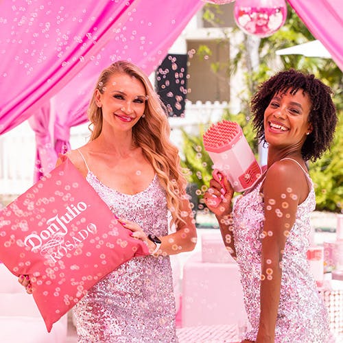 Two women at a Don Julio Rosado event. One Woman is holding a pink Don Julio Rosado pillow while the other is blowing bubbles using a bubble gun.